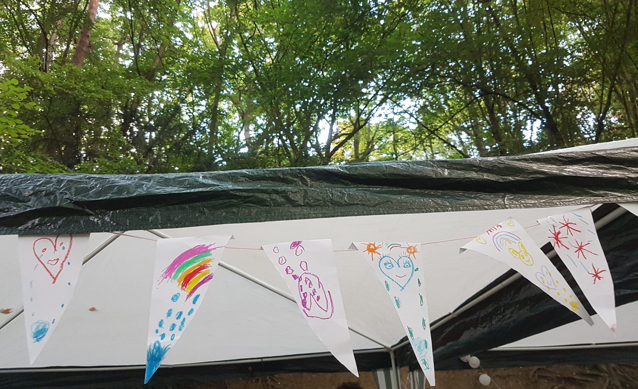 Im Wald an einem Pavillon Dach, das mit einer Plane abgedeckt ist, hängen selbstgemachte Fähnchen mit Kinderzeichnungen.