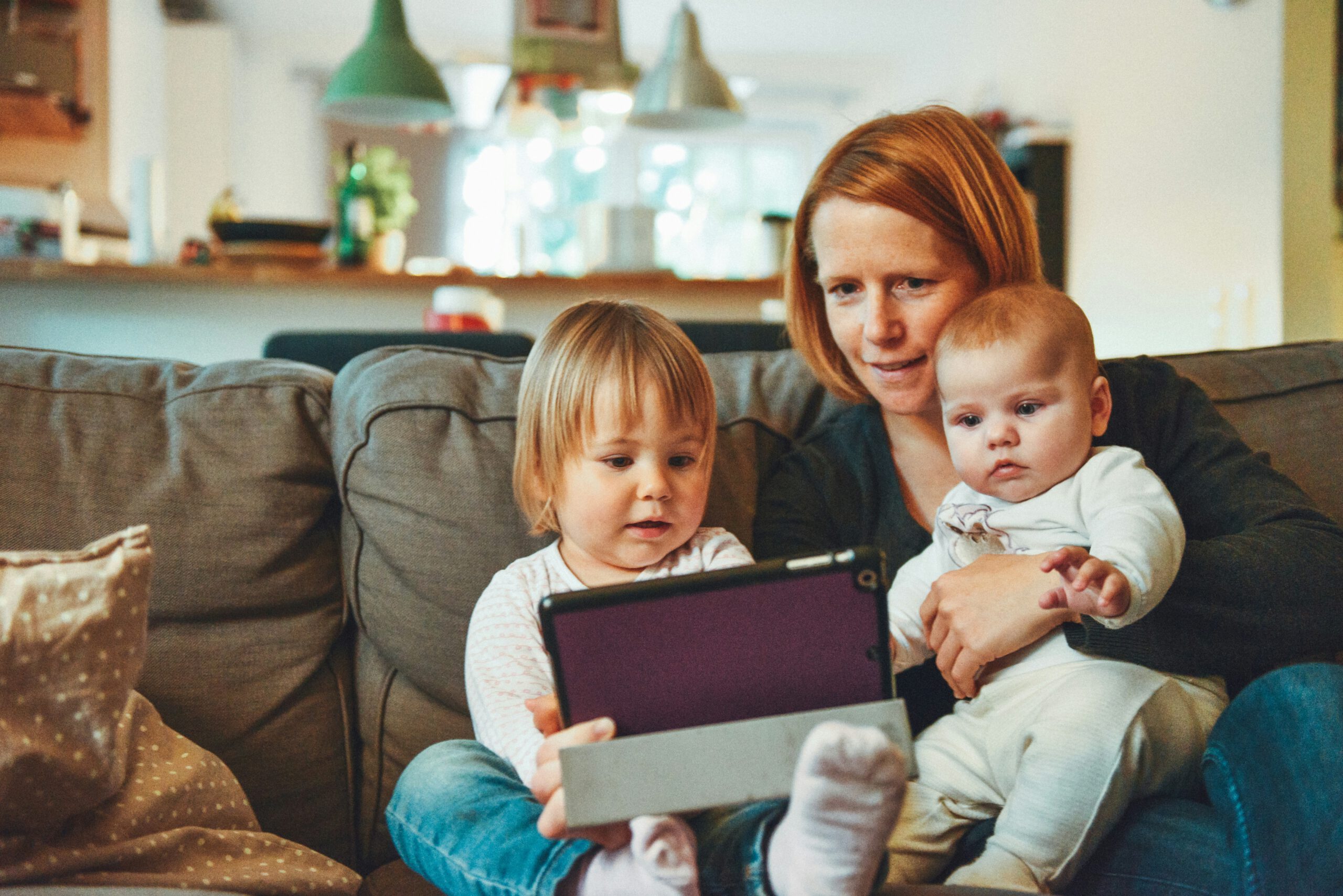 Mutter sitzt mit zwei kleinen Kindern auf dem Sofa und schaut mit ihnen auf ein Tablet.